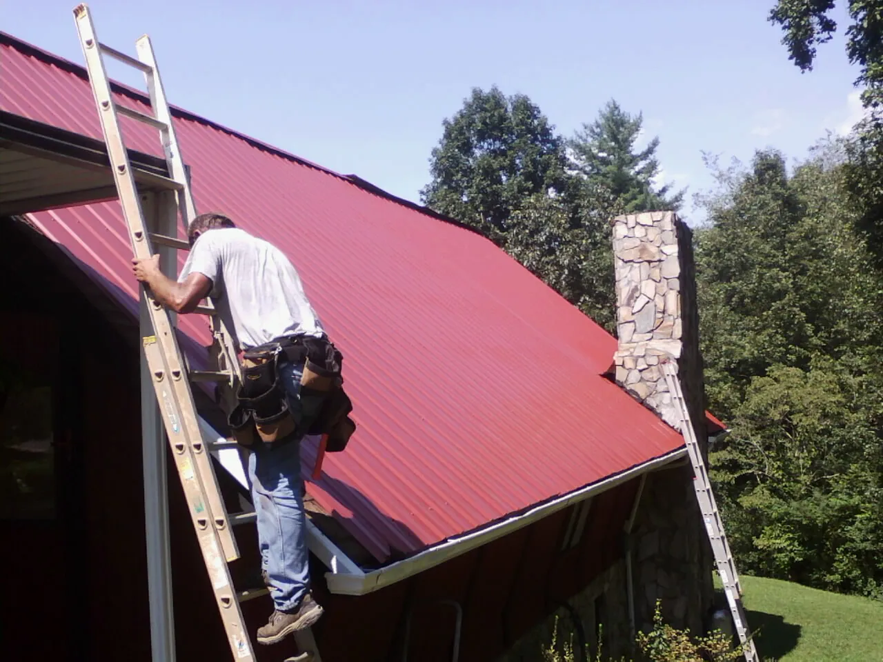 Roofer on a ladder beside a red metal roof and stone chimney — Soffit & Fascia Repair by Roofing Brothers in Buckhead